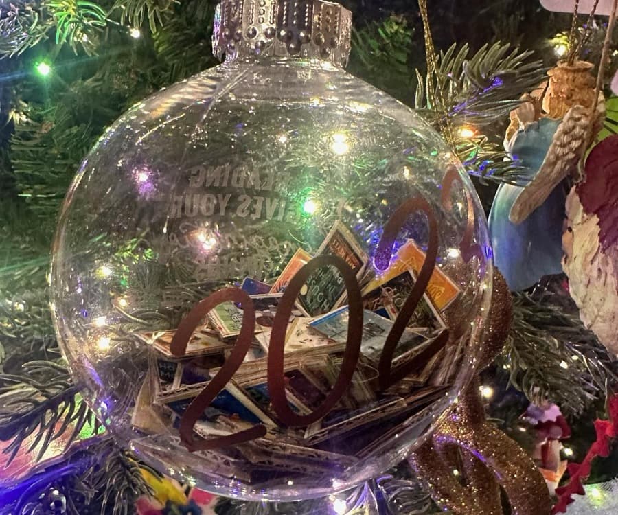 A clear glass fillable ornament packed with miniature book covers and labeled with a decorative year numeral, hanging on a Christmas tree lit with colorful lights.