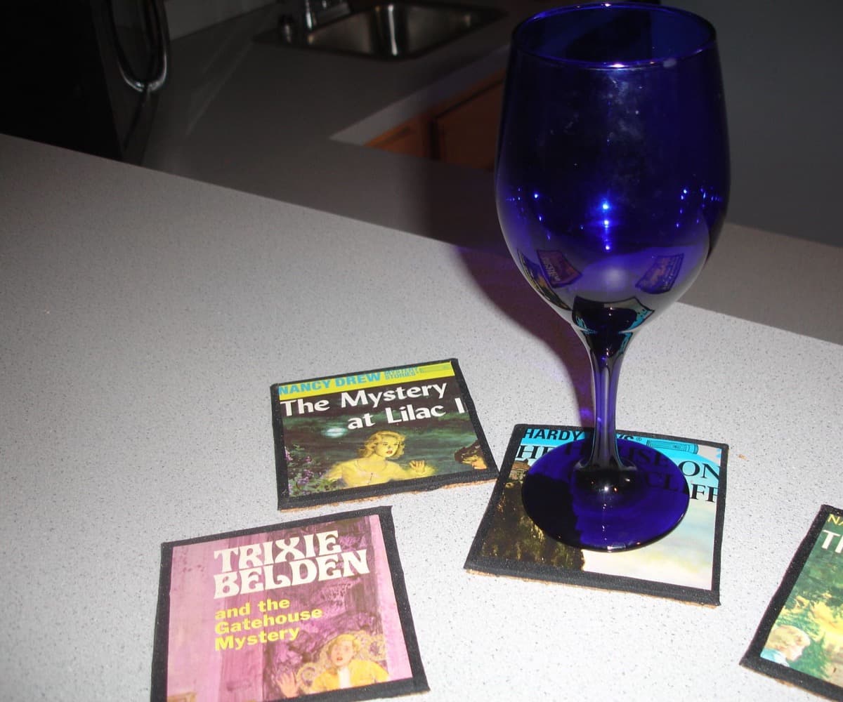 Four coasters made from vintage Nancy Drew, Hardy Boys and Trixie Belden book covers arranged on a kitchen counter, with a deep blue wine glass resting on one of them.
