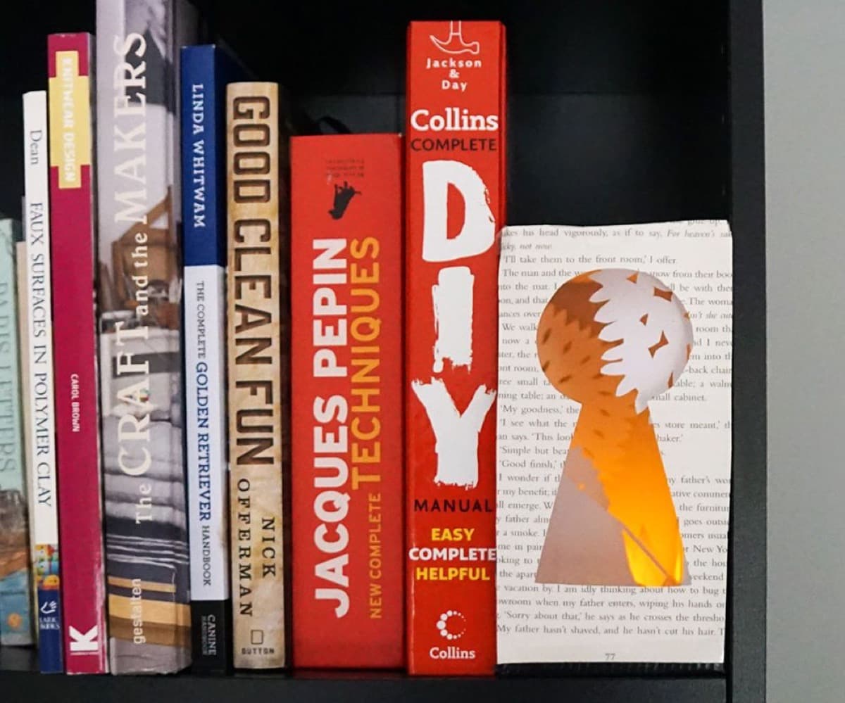 A papercut keyhole shape cut from book pages glowing warm orange from a hidden tealight, tucked between a row of real books on a dark shelf.