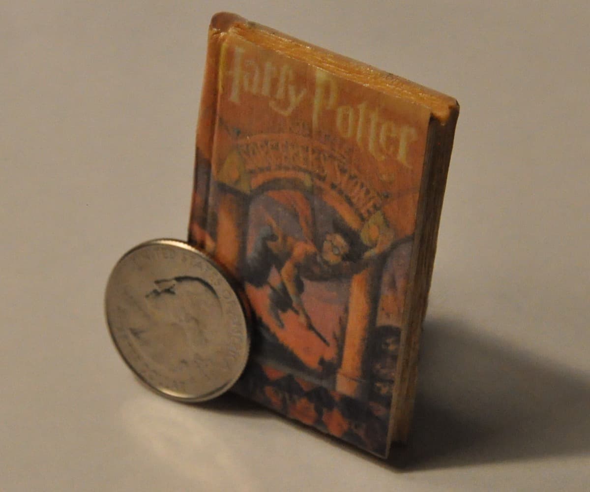 A miniature hand-bound book with a faded Harry Potter-style cover stood on end beside a US quarter for scale on a plain backdrop.