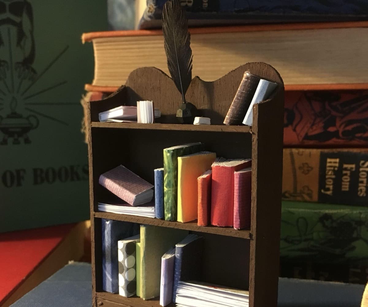 A small hand-made wooden bookcase filled with tiny colorful handmade books with a black quill pen tucked into the top shelf, photographed against a backdrop of stacked vintage hardcovers.