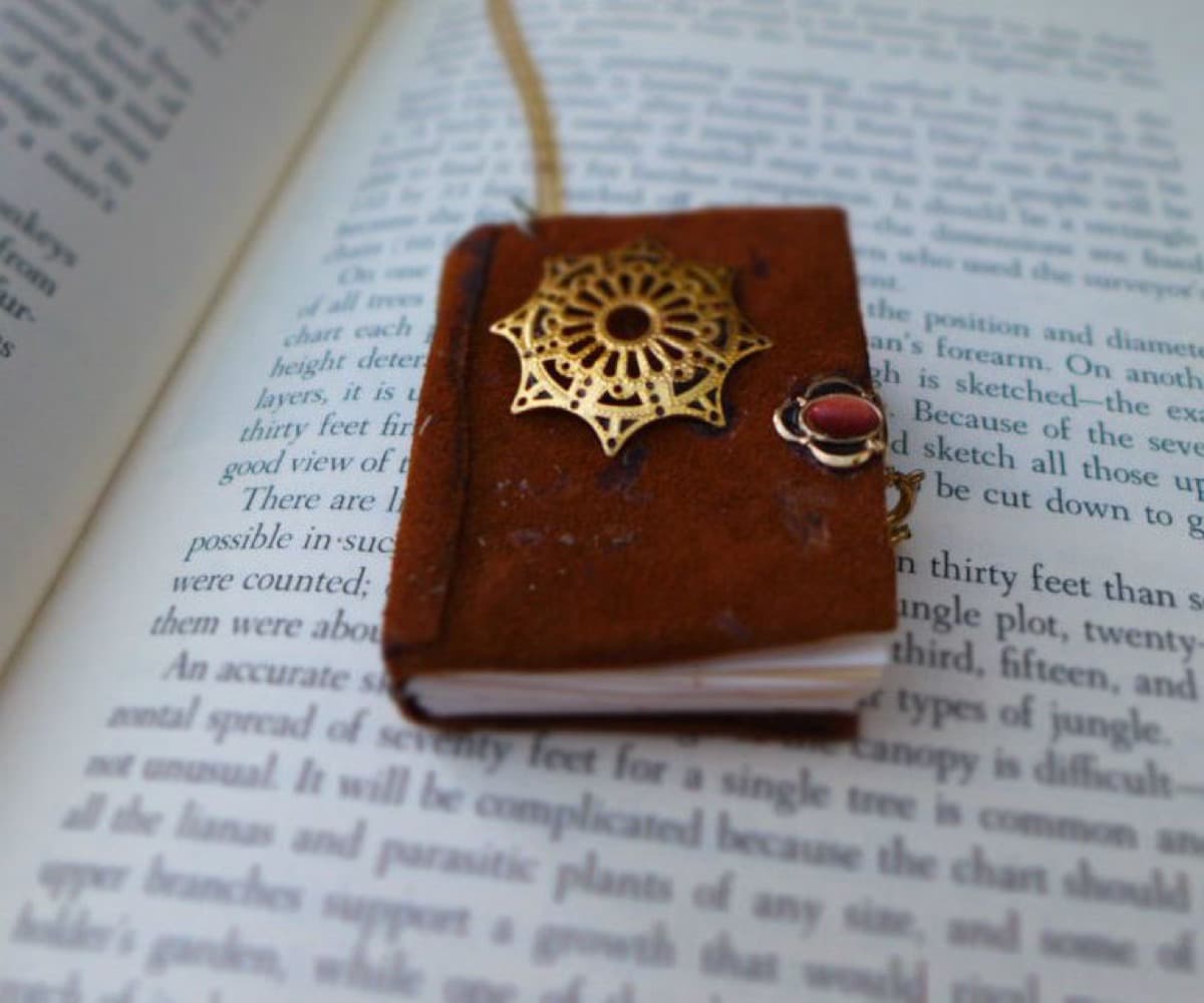A small brown leather-bound mini book pendant with a gold filigree ornament and a red gem clasp, resting open-face on the printed page of a full-size book.